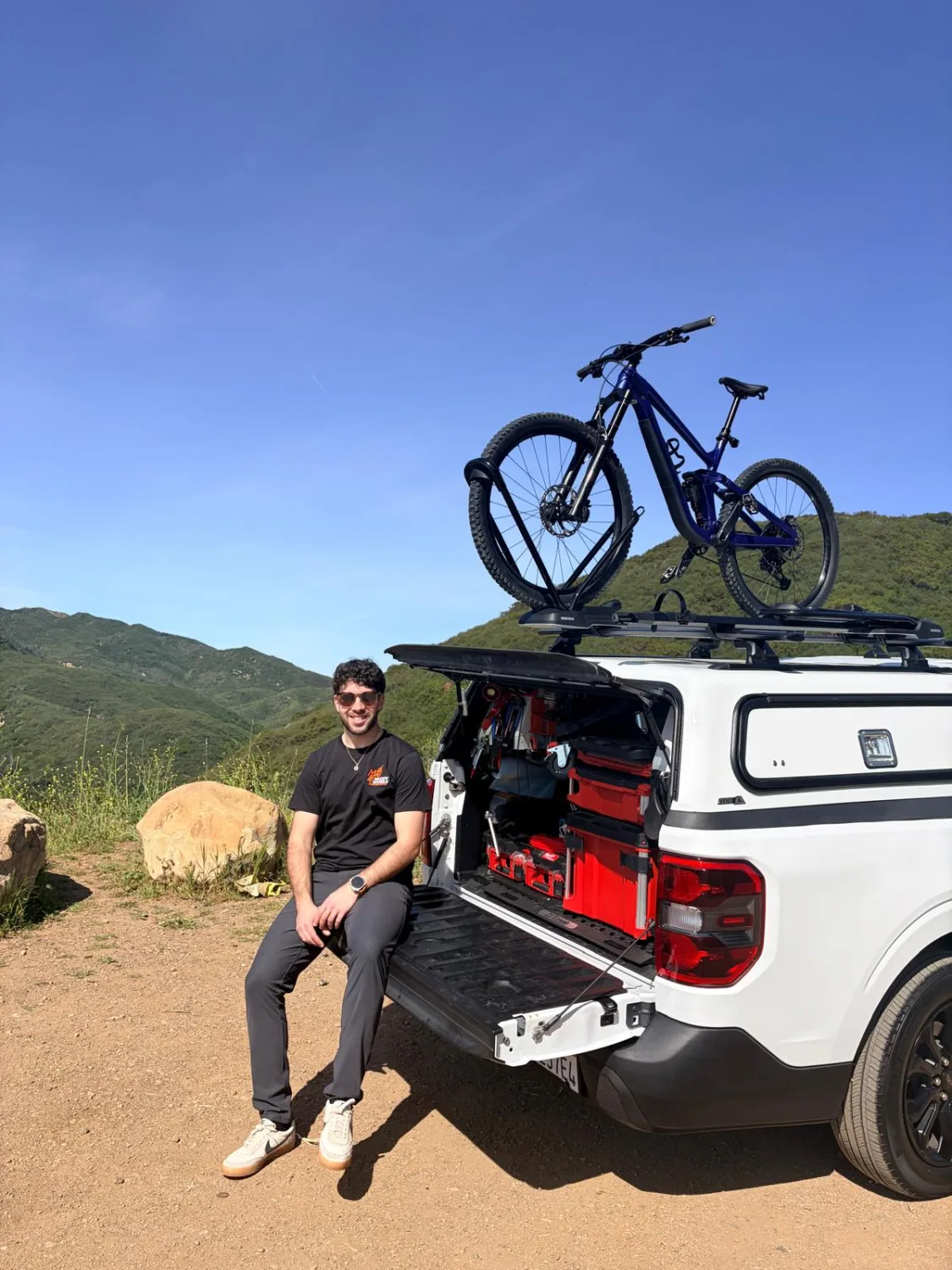 Shlomi Bar Lev of Muddy Wheels on the tailgate of his mobile workshop truck, bike on the roof rack, Los Angeles hills behind